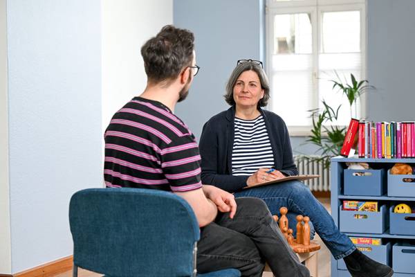 Eine Frau berät einen Mann in einem Raum der Evangelischen Beratungsstelle für Erziehungs-, Ehe- und Lebensfragen in Mülheim an der Ruhr. Die Frau sitzt mit dem Gesicht zur Kamera dem Mann gegenüber, der mit dem Rücken zur Kamera sitzt. Sie hat kinnlanges graues Haar und eine Lesebrille nach oben auf den Kopf geschoben. Sie trägt ein schwarz-weiß gestreiftes Oberteil mit einer schwarzen Strickjacke darüber. Sie hat ein Klemmbrett in der Hand und redet gerade. Der Mann trägt ein pink-schwarz gestreiftes T-Shirt und man erkennt, dass er eine Brille und einen Bart hat.
In dem Raum sind rechts Bücher in einem Regal zu erkennen, sowie diverse Utensilien für Beratungen - zum Beispiel Holzfiguren, ein Plüschtier und Spielzeug.
Im Hintergrund erkennt man eine Pflanze und eine Sitzgruppe mit Kinderstühlen.